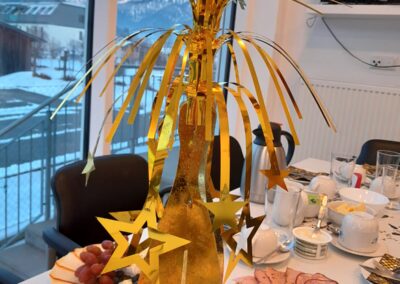 Goldene Tischdekoration mit Sternen auf einem gedeckten Frühstückstisch; Aufschnitt, Käse und Lachs, verschneite Berge durch Fenster im Hintergrund.