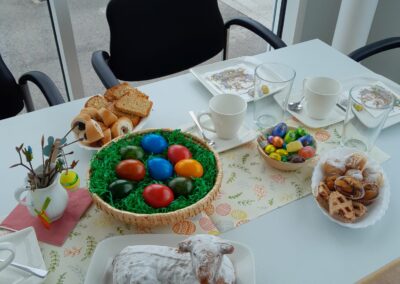 Tisch mit Osterdekoration: Korb mit bunten Eiern, Lammkuchen mit Puderzucker, Gebäck, Schokoladeneiern und Kaffeetassen.