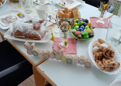 Gedeckter Tisch mit Ostergebäck, Schale bunter Eier und Holzschriftzug 'FROHE OSTERN'.