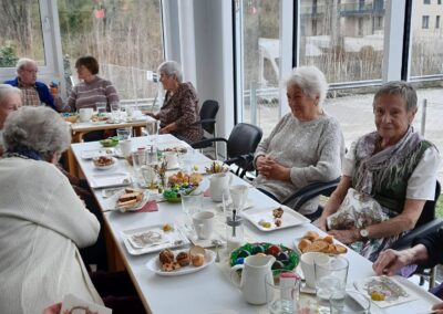 Ältere Personen an einem gedeckten Tisch mit Osterdekoration, bunten Eiern, Süßigkeiten und Getränken in einem hellen Raum mit großen Fenstern.