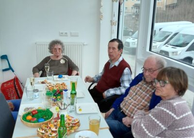 Vier ältere Personen an einem gedeckten Tisch mit Osterdekoration, bunten Eiern und Snacks in einem hellen Raum mit Fenster.