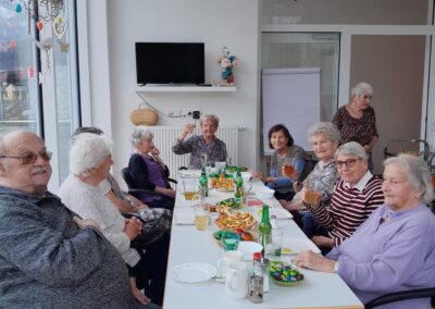 Mehrere ältere Menschen sitzen an einem langen Tisch mit Getränken, Snacks und Osterdekoration in einem hellen Raum.