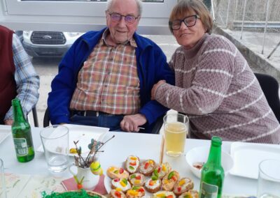 Zwei Personen sitzen nebeneinander an einem Tisch mit Osterdekoration, bunten Eiern und Häppchen.