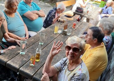 Ältere Menschen sitzen an langen Holztischen im Freien unter Bäumen; Frau winkt in die Kamera; Getränke und Tischschild mit der Zahl 13 sichtbar.