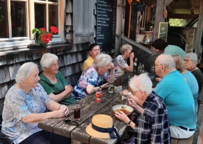 Außensitzbereich eines Gasthauses, mehrere ältere Personen sitzen an rustikalem Holztisch, essen (Suppe) und trinken; an der Hauswand Kreidetafel mit Schrift "Zipfer" und Speisekarte, Blumen am Fenster.