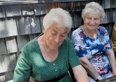 Zwei ältere Frauen an einem Holztisch; die linke in grün hält eine Pfanne mit zerkleinertem Pfannkuchengericht, im Hintergrund ein Fensterbrett mit roter Geranie.