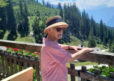 Person mit Sonnenhut und Sonnenbrille lehnt an einem Holzgeländer; im Hintergrund bewaldete Berge und blauer Himmel.