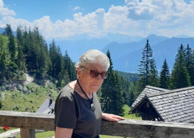 Ältere Frau mit Sonnenbrille steht an einem Holzgeländer auf einer Terrasse; im Hintergrund Tannen, Holzhütten und ein Bergpanorama unter blauem Himmel mit Wolken.