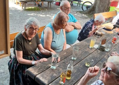 Ältere Personen sitzen an einem langen Holztisch im Freien; Gläser und Flaschen auf dem Tisch; Sonnenschirm mit Schriftzug ‚Gösser‘ sichtbar.