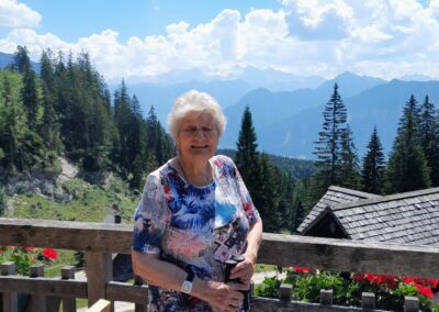 Ältere Frau steht auf einer Holzterrasse in den Bergen, lächelt und hält Wanderstöcke; rote Blumen am Geländer, Tannen und Bergketten im Hintergrund.