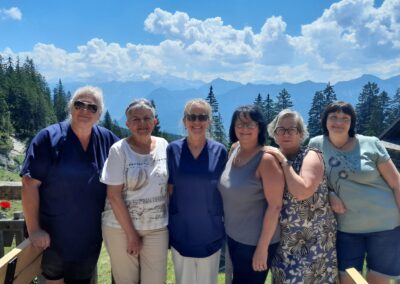 Sechs Frauen stehen dicht nebeneinander auf einer Holzterrasse, im Hintergrund Nadelbäume und bergiges Panorama unter blauem Himmel mit Wolken.