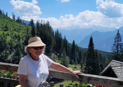 Person mit Sonnenhut und Sonnenbrille lehnt an einem Holzgeländer; dahinter Tannen, Bergkette und blauer Himmel mit Wolken
