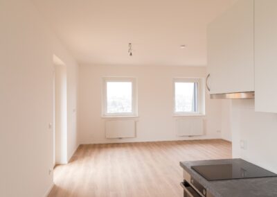 Leerer, heller Wohnraum mit zwei Fenstern, Heizkörpern, Küchenzeile und freihängender Deckenlampe