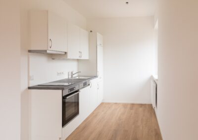 Schmale weiße Küchenzeile mit Spüle, Cerankochfeld und Einbauofen, helle Wände und Holzfußboden
