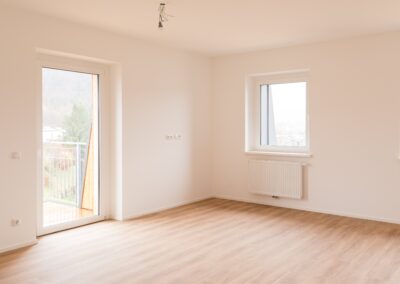 Leerer Innenraum mit hellem Holzboden, Fenster, Balkontür und Heizkörper.