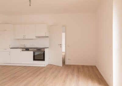 Leerer Raum mit weißer Küchenzeile, Spüle, Einbauofen und offen stehender Tür.