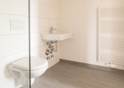 Helles, modernes Badezimmer mit wandhängendem WC, kleinem Wandwaschbecken und Handtuchheizkörper; Spülplatte mit zwei Kreisen sichtbar.
