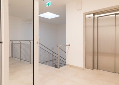 Heller Innenflur mit geschlossener Aufzugstür rechts, Glastür links, Sicht auf Treppe und grünem Notausgangsschild an der Decke.