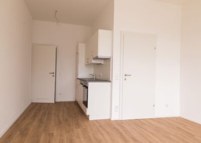 Innenaufnahme eines leeren Wohnraums mit kleiner Einbauküche, weißen Wänden, Holzfußboden und freiliegenden Kabeln an der Decke.