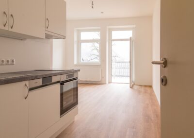Leere, helle Wohnungsküche mit Arbeitsplatte, Einbaubackofen, Hängeschränken und offener Balkontür.