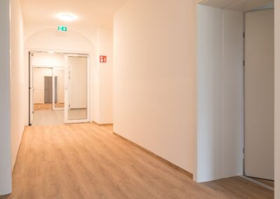 Heller leerer Flur mit Holzfußboden, Glastür, grünem Notausgangsschild und rotem Feuerlöschersymbol.