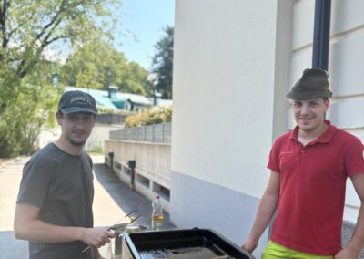 Zwei Männer stehen neben einem Tischgrill mit bratenden Würstchen; im Vordergrund mehrere Teller, im Hintergrund Bäume und eine Hausfassade; Kappe mit Schrift 'LOS ANGELES' sichtbar.