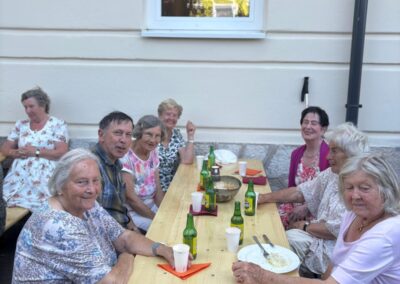 Acht ältere Personen sitzen an einem langen Holztisch draußen vor einer hellen Hauswand; auf dem Tisch stehen grüne Flaschen, Pappbecher und Teller; im Fenster spiegelt sich ein gelbes Haus.