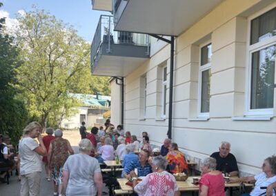 Mehrere ältere Menschen sitzen an aufgestellten Holztischen und -bänken entlang einer Hausfassade; auf den Tischen Getränke und Snacks; einige Personen nutzen Gehhilfen.