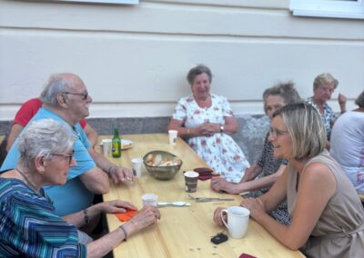 Mehrere ältere Personen sitzen an einem langen Holztisch vor einer Hausfassade; auf dem Tisch Tassen, eine Schüssel mit Brot und Besteck.