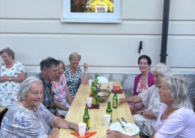 Ältere Personen sitzen an einem langen Holztisch draußen neben einer Hauswand; Getränke, Becher und Teller auf dem Tisch; Fenster mit Spiegelung eines gelben Hauses.