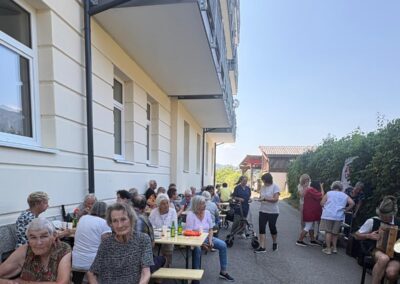 Ältere Menschen sitzen an langen Holztischen neben einem Wohngebäude, trinken Kaffee und essen Kuchen; einige stehen, eine Person spielt Akkordeon.