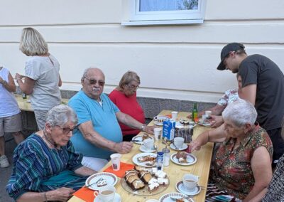 Mehrere ältere Personen und ein junger Mann an einem Holztisch im Freien, auf dem Kuchen, Kaffeetassen und ein Milchkarton mit der Aufschrift 'Vollmilch' stehen.