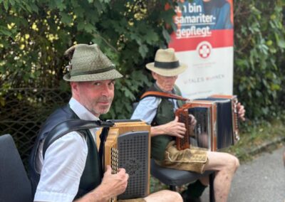 Zwei Männer sitzen im Garten und spielen diatonische Akkordeons, sie tragen traditionelle Tracht (Hut, Lederhosen). Im Hintergrund ein Roll‑up mit dem Schriftzug "Ich bin samaritergepflegt".
