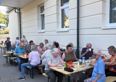 Ältere Menschen sitzen an langen Tischen draußen neben einem Haus und essen.