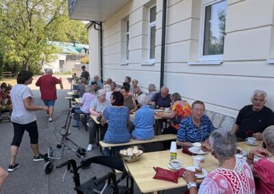 Ältere Menschen sitzen an langen Holztischen vor einem Wohngebäude, trinken Kaffee und essen Kuchen; ein Rollator im Vordergrund.